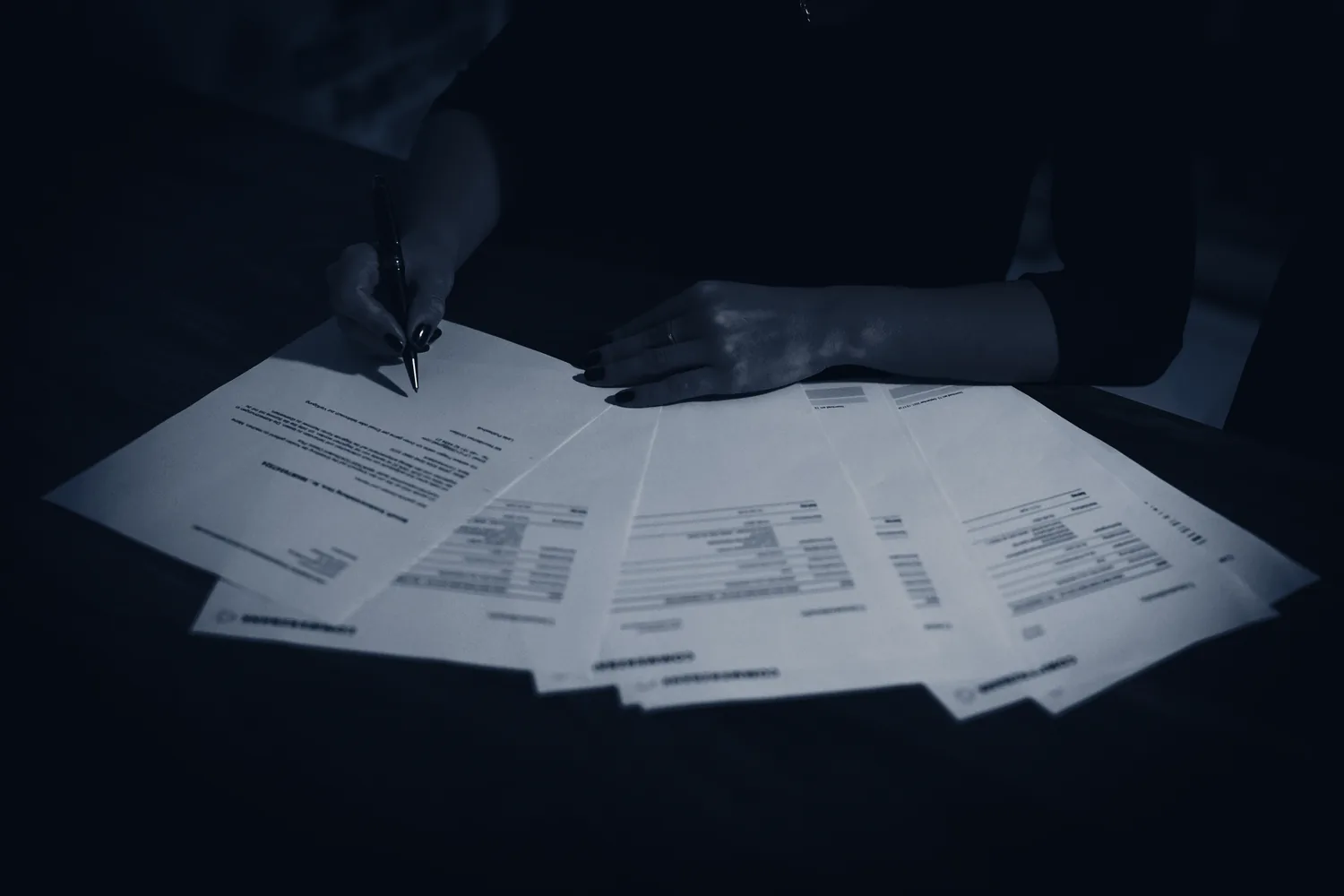 Hands annotating documents laid out across a dark desk